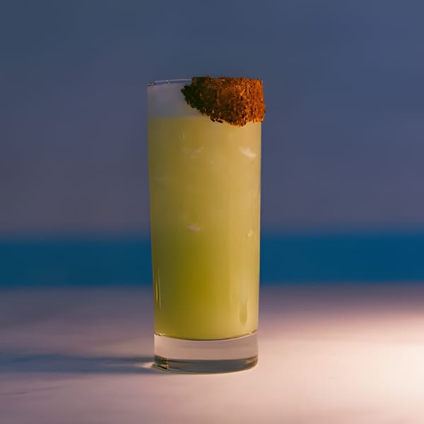 Tall glass of light green cocktail with a brown, textured rim against a blue background.