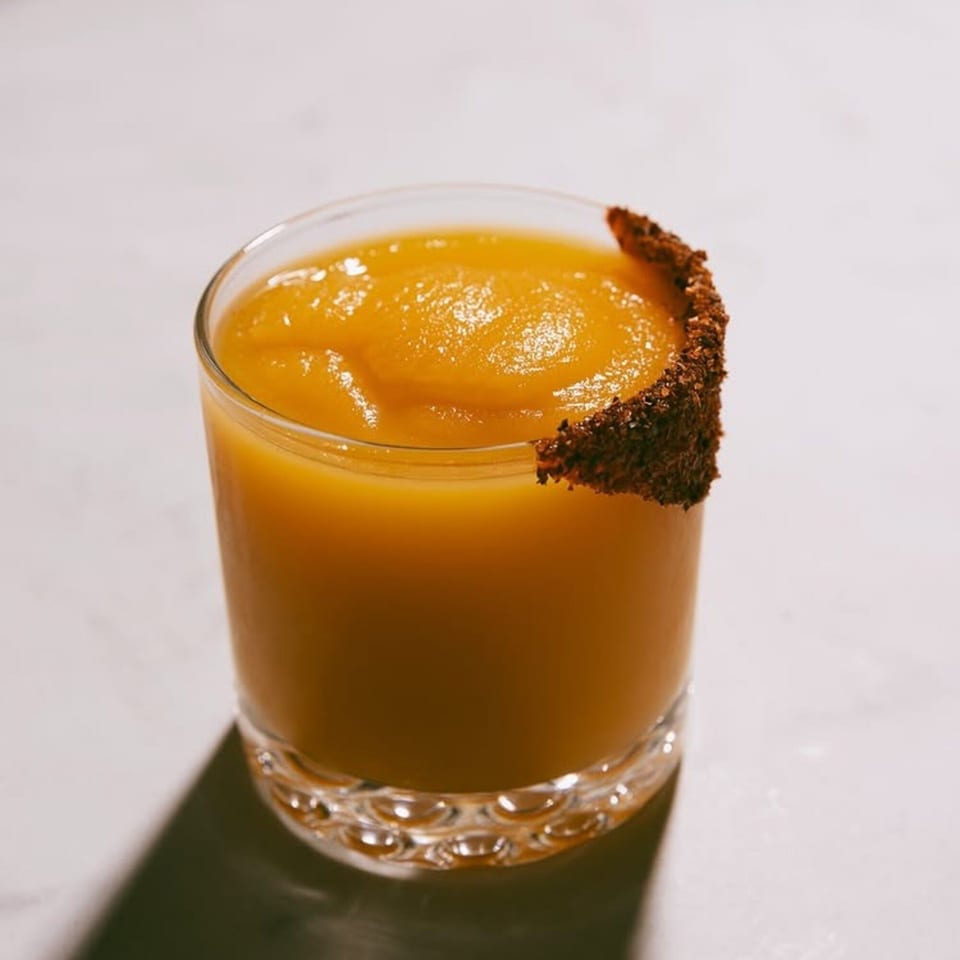 A vibrant orange frozen cocktail in a glass with a dark, spiced rim, casting a shadow.