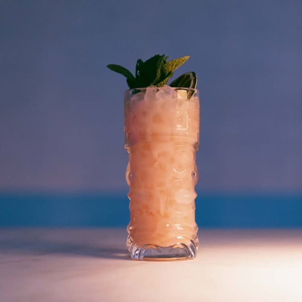 A tall, carved tiki glass filled with a light pink cocktail, crushed ice, and mint garnish.