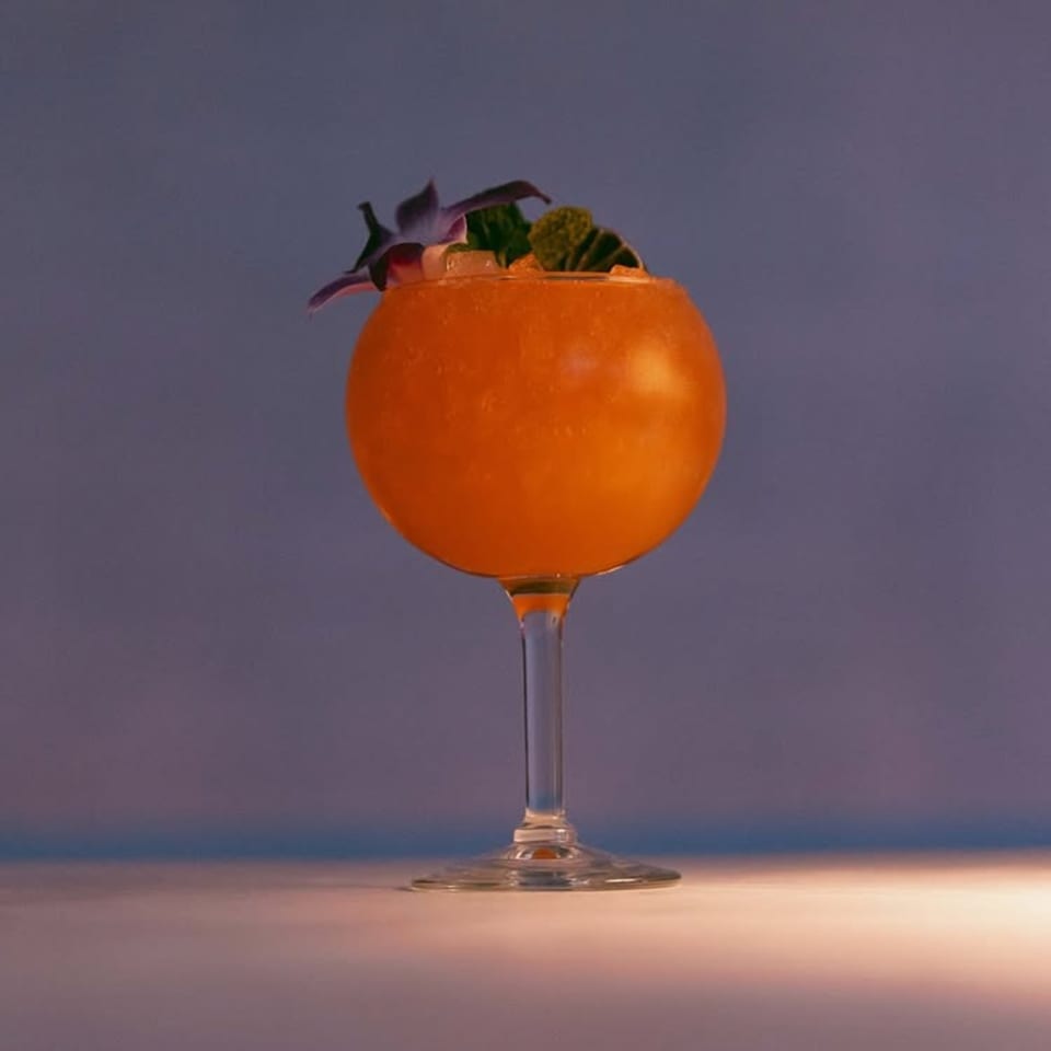 A vibrant orange cocktail in a stemmed glass, garnished with a purple flower and green leaves.