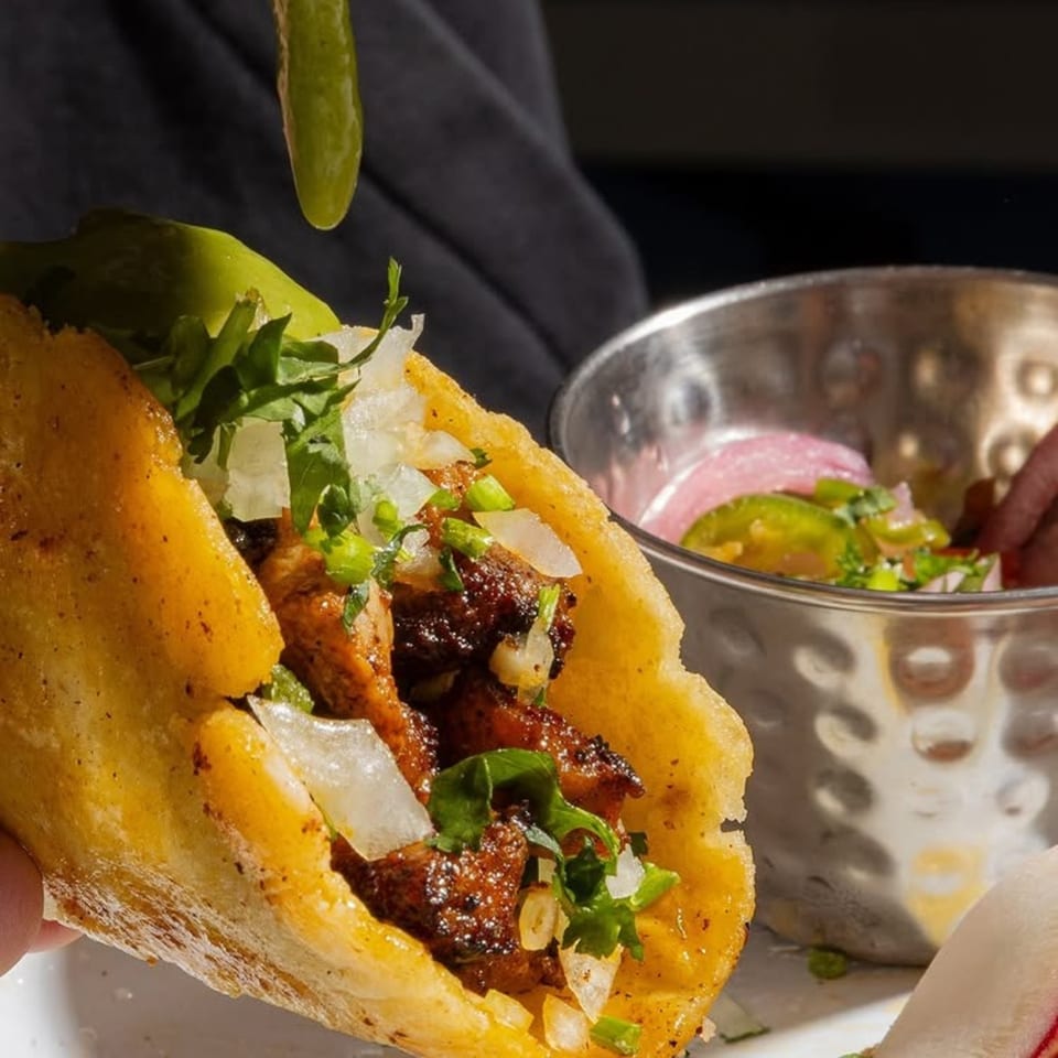 A hand holds a crispy taco filled with meat, onions, and cilantro, with a side of salsa.