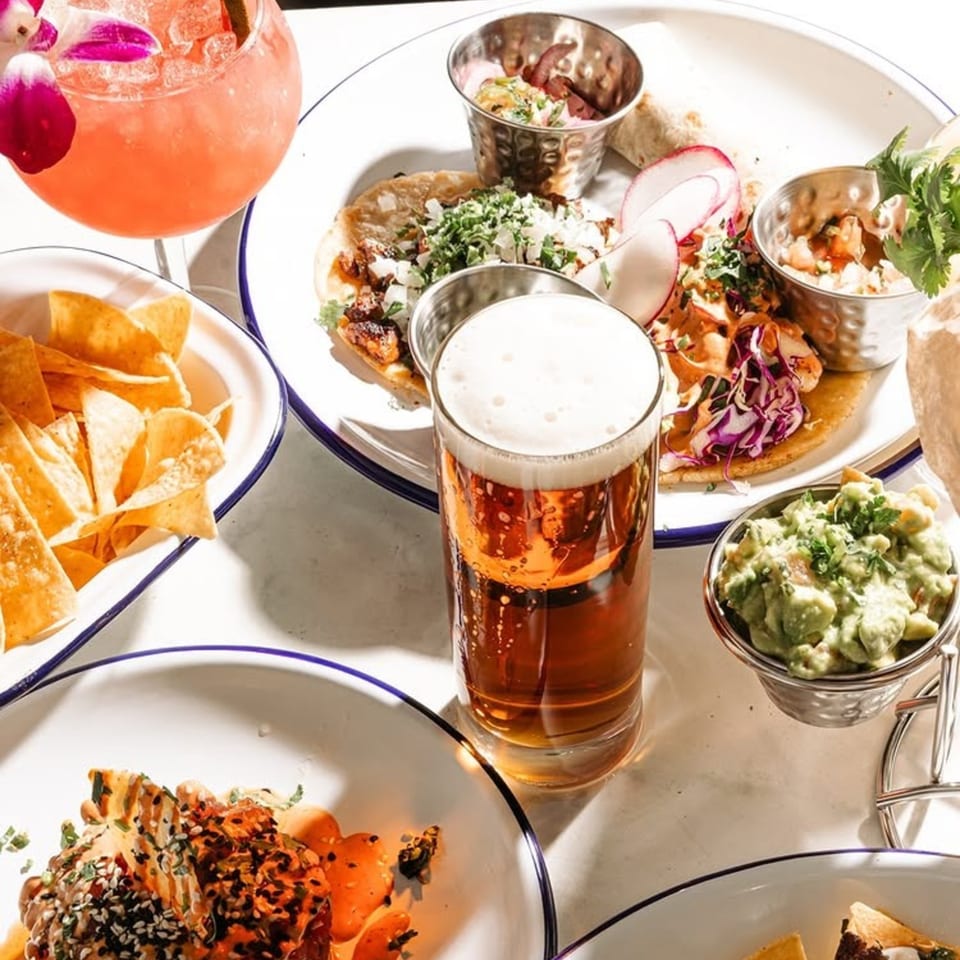 A table spread with tacos, guacamole, chips, a beer, and a pink cocktail with an orchid.