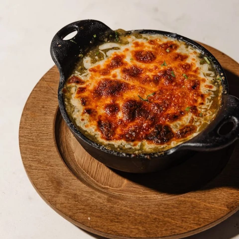 A cast iron skillet of bubbling green chili queso with melted, browned cheese on a wooden plate.