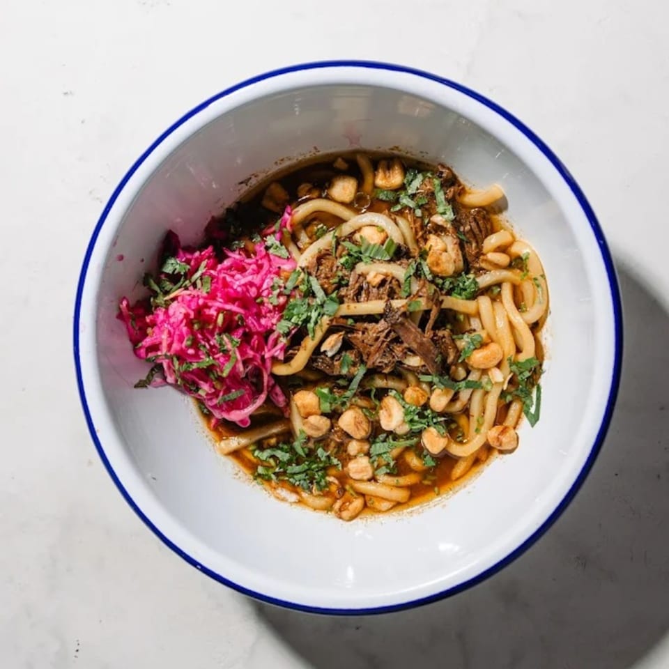 A bowl of udon noodles with shredded meat, peanuts, cilantro, and pickled red cabbage.