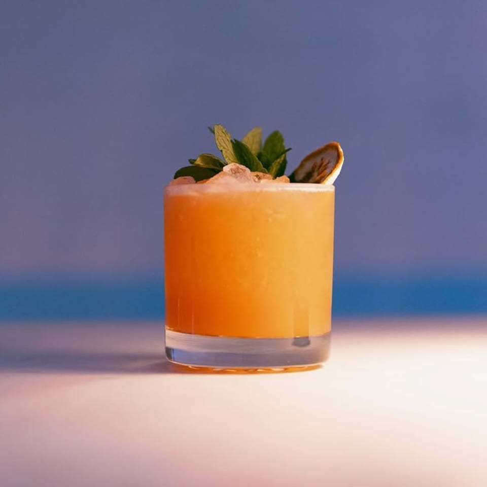A vibrant orange cocktail, garnished with fresh mint and a dried citrus slice, against a blue background.