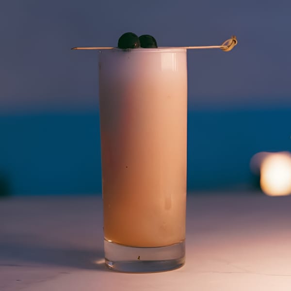 A tall, light-colored cocktail with two dark berries on a skewer, against a blue background.